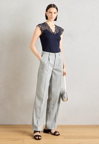 Haut bleu marine avec des manches en dentelle, associé à un pantalon large à rayures grises claires et des sandales plates noires. Mannequin tenant un sac argenté.