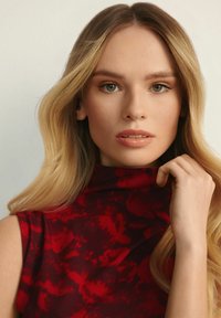 Czerwony bezrękawnik z wysokim dekoltem, ozdobiony kwiatowym wzorem. Gładka tekstura materiału oraz modelka poprawiająca kołnierz, z delikatnymi, falującymi włosami.