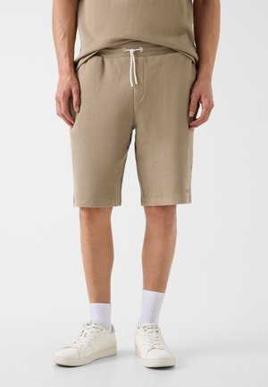 Mann trägt beige strukturierte Shorts mit Kordelzug, weiße Kniesocken und weiße Turnschuhe vor einfarbigem Hintergrund.