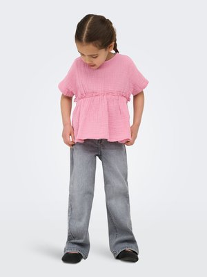 Ragazza giovane con capelli trecciati che indossa una camicetta rosa larga, jeans grigi a gamba larga e scarpe nere, guarda verso il basso il suo outfit.