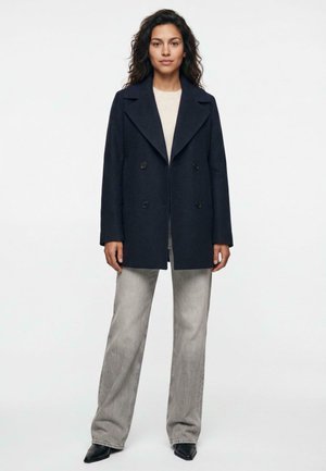 Woman wearing a dark navy double-breasted coat, beige top, light gray wide-leg jeans, and black pointed shoes standing against a white background.