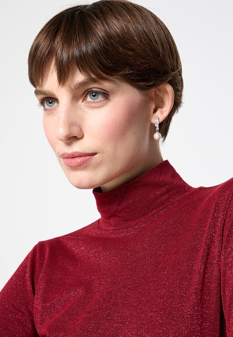 Rotes, eng anliegendes Rollkragenoberteil mit glitzernder Textur. Modell trägt hängende Ohrringe mit einer Perle und einem Kristallakzent.
