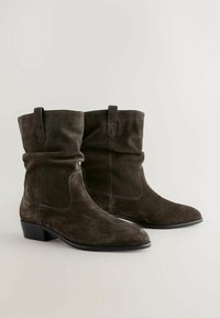 Bottines en suède marron avec un design décontracté, bouts pointus et un petit talon empilé. Présente des coutures discrètes et des tirants sur la tige.