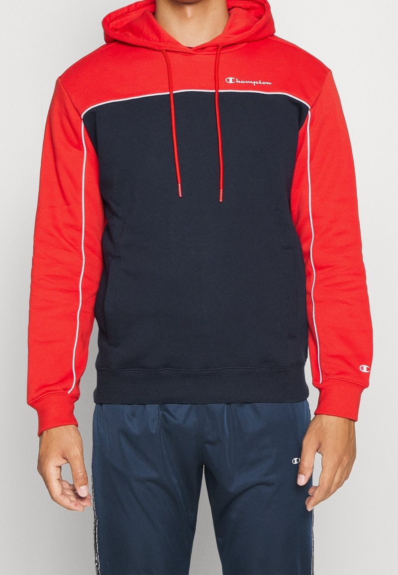 Homme portant un sweat à capuche Champion rouge et bleu marine avec des cordons rouges et un pantalon de survêtement Champion bleu marine avec des bandes blanches sur les côtés.