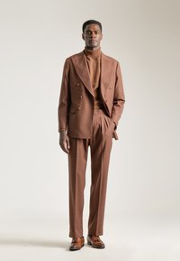 Chaqueta de traje marrón de doble botonadura con botones dorados, combinada con un suéter de cuello alto marrón ajustado y pantalones a juego. Unos zapatos de cuero marrón completan el conjunto.