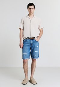 Un giovane uomo sta in piedi con una mano in tasca, indossando una camicia beige a maniche corte, pantaloni corti in denim strappati con cintura e scarpe slip-on beige.