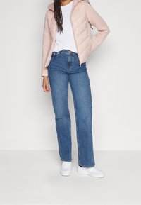 Ljusrosa quiltad jacka med huva över en vit t-shirt, kombinerad med högmidjade blå jeans och vita sneakers.