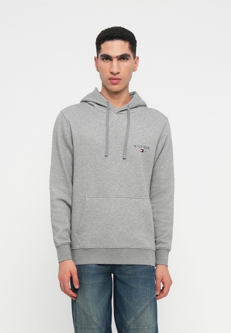 Gray cotton hoodie with a front pocket, drawstring, and logo detail on the chest, paired with blue jeans featuring a textured pattern.