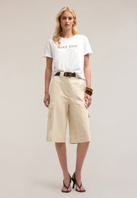 Vit t-shirt med guldtexten "MAKE TODAY", kombinerad med beige cargo-shorts med två sidofickor och ett mörkt bälte. Bruna armband.