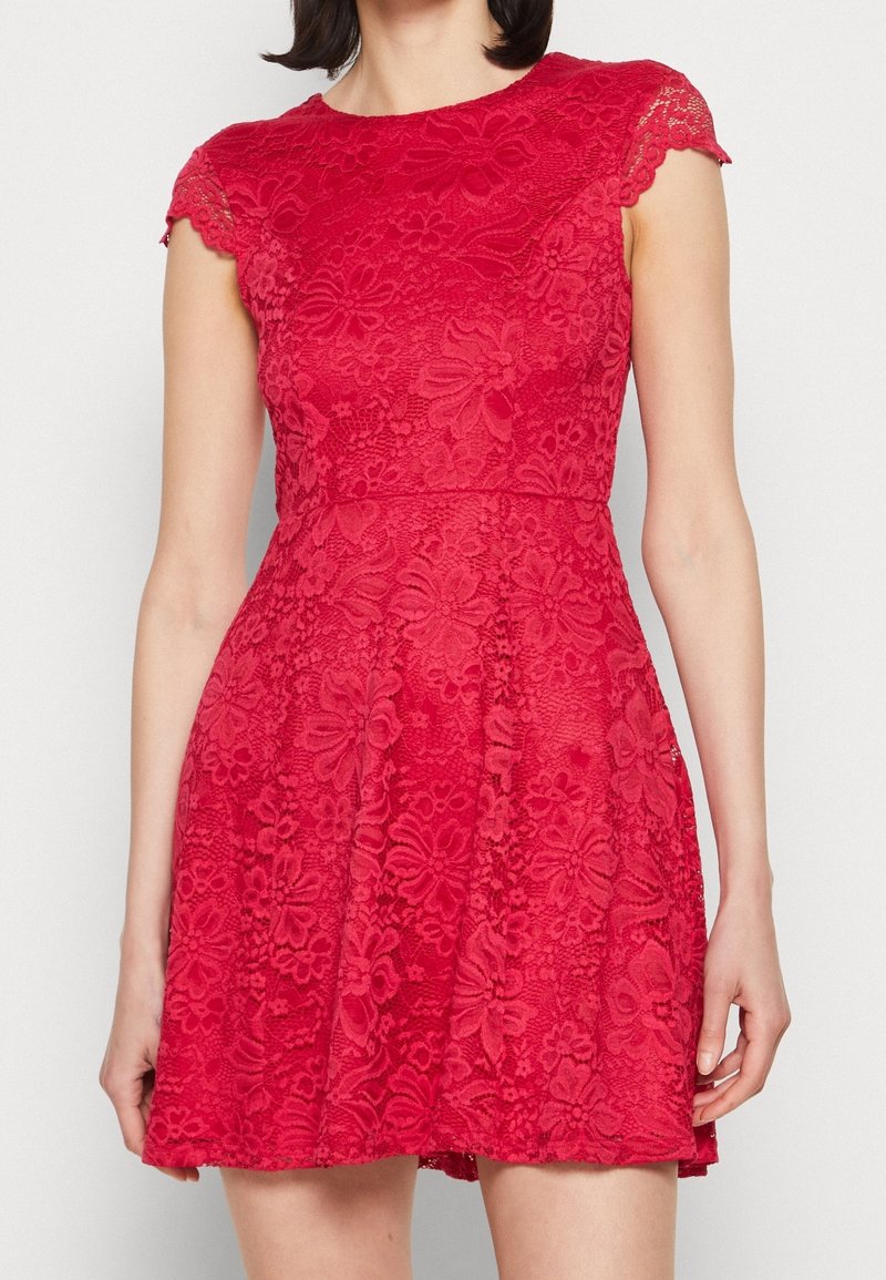 Femme portant une robe rouge en dentelle à manches courtes avec des motifs floraux et une taille cintrée, debout devant un fond clair uni.