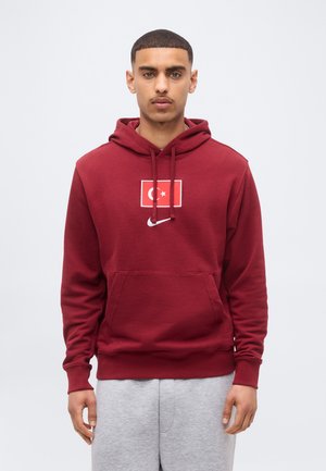 Mlad muškarac stoji, nosi tamnocrvenu duksericu s kapuljačom s turskom zastavom i bijelim Nike logotipom, u kombinaciji sa svijetlosivim trenirkama, neutralna siva pozadina.