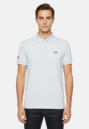 Giovane uomo in piedi con postura eretta che indossa una polo bianca a maniche corte con piccoli loghi rossi sul petto e sulla manica sinistra.