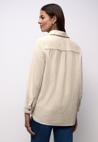 Beżowa teksturowana koszula z kołnierzem, długimi rękawami oraz mankietami na guziki, o luźnym kroju i detalem na plecach.