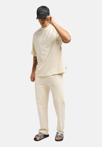 Beige bomulls oversized t-skjorte, matchende joggebukser og svart caps. Sklisandaler har et slangeskinnmønster med svart såle.