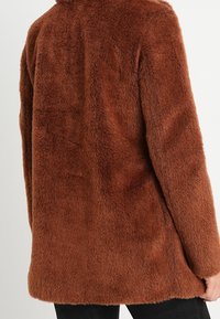 Brun fuskpälsjacka med en mjuk, texturerad yta. Halvlång design, långa ärmar och rund halsringning. Inga synliga knäppningar.