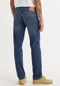 Blaue Jeans aus Denim mit geradem Bein, ausgestattet mit Gesäßtaschen, einem Lederpatch an der Taille und abgedrifteter Textur. Kombiniert mit braunen Schuhen.