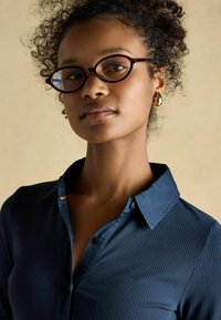 Dark blue ribbed shirt with a button-up collar, featuring a subtle accent at the neck. Round glasses and gold hoop earrings are worn.