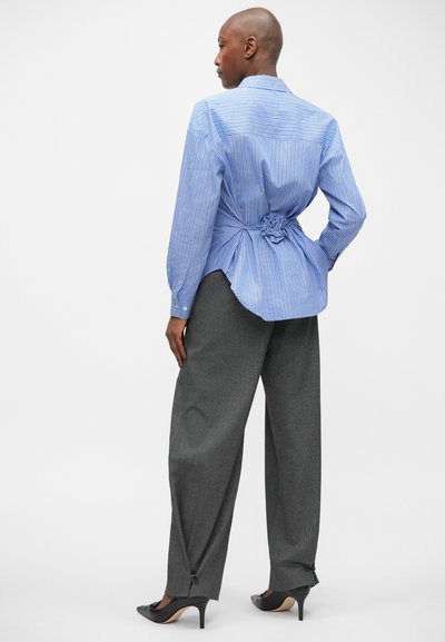 Blauw-witte gestreepte blouse met lange mouwen, aan de achterkant geknoopt; gecombineerd met een grijze broek met wijde pijpen en zwarte hakken.