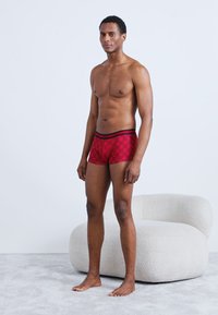 Κόκκινα ανδρικά μποξεράκια με γεωμετρικό μαύρο σχέδιο και μαύρη ζώνη στη μέση. Το μοντέλο στέκεται πάνω σε ανοιχτόχρωμο χαλί δίπλα σε μια καρέκλα με υφή.