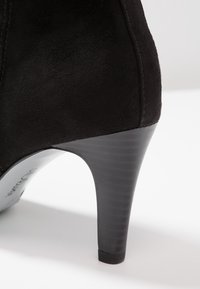 Black suede high-heeled shoe with a tapered heel. Textured heel design with a smooth interior sole. Side seam detail visible.