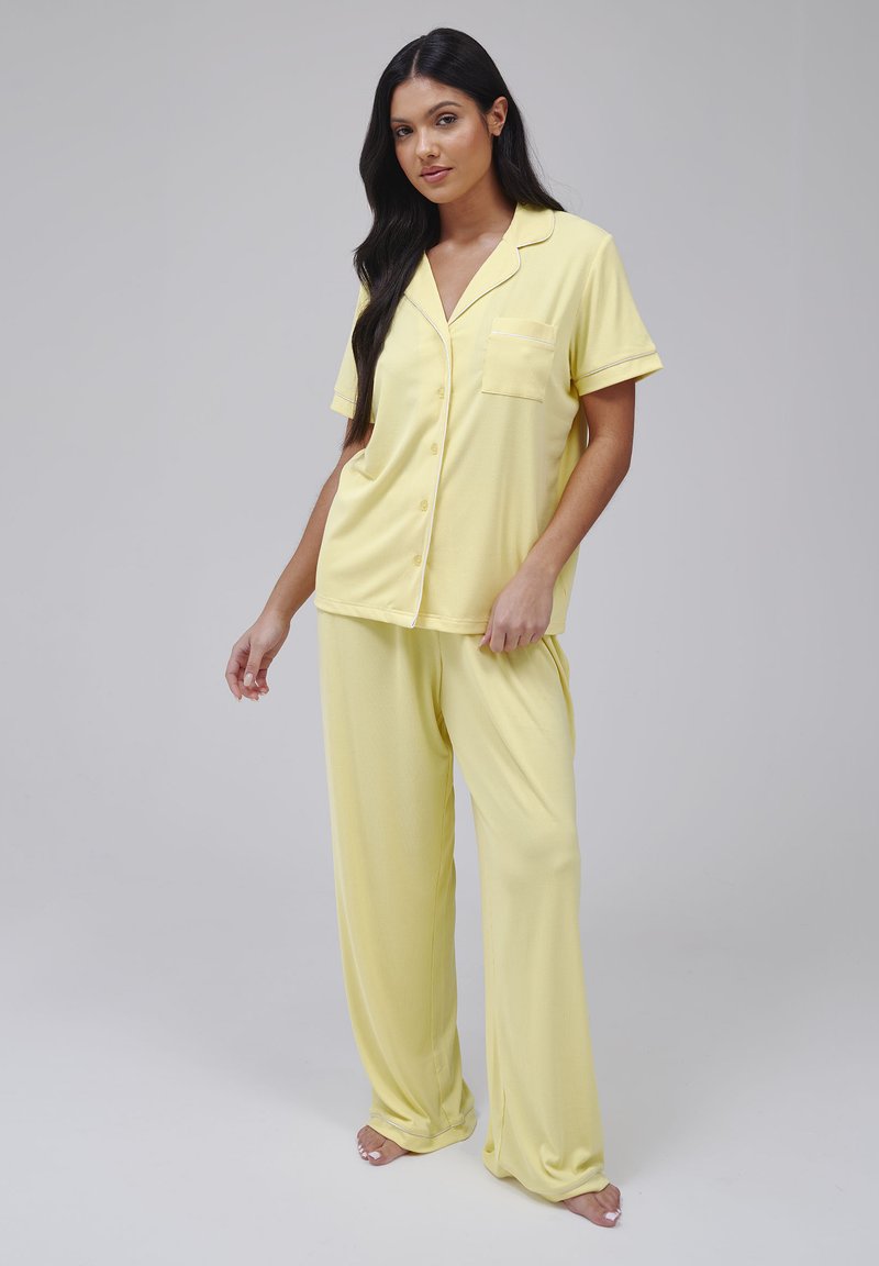 Pyjamapak in zachte, gele stof. Korte mouw shirt met een zakje en contrasterende bies. Loszittende broek met een elastische tailleband.