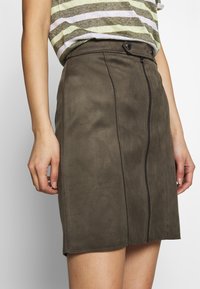 Olivgrüner Wildledrock mit einem Frontreißverschluss, zwei Knopfdetails am Bund und einer schlanken Silhouette. Weicher Stoff und taillierte Passform.