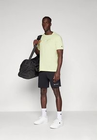 Must kott käes; seljas helekollane T-särk logoga, mustad lühikesed püksid ja valged tennised; seisab ühetoonilisel taustal.