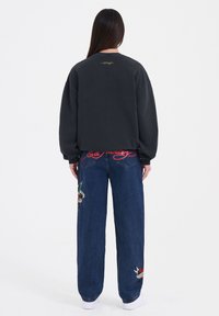 Czarna, oversize bluza z ribbowymi mankietami, z małym, zielonym logo z tyłu. Połączona z szerokimi, niebieskimi dżinsami zdobionymi kolorowymi haftami.