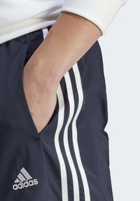 Námořnické sportovní šortky s třemi bílými pruhy po straně a logem Adidas nalevo. Tkanina vypadá hladce a je lehká.