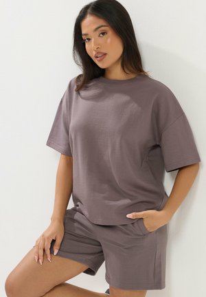 Mujer apoyada contra una pared, vistiendo una camiseta holgada de color malva y pantalones cortos a juego, con una mano en el bolsillo y la otra descansando sobre su muslo.