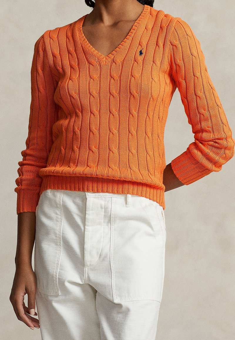 Femme portant un pull en maille torsadée orange vif à col en V et un pantalon blanc taille haute, une main posée sur sa hanche.