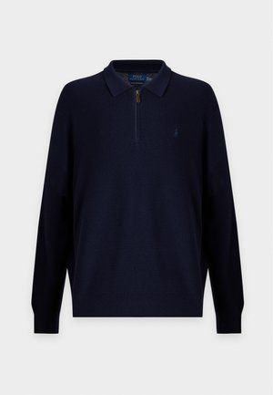 Jeune homme aux cheveux bouclés portant un polo bleu foncé à manches longues, un jean bleu clair et des baskets blanches, debout avec une main dans la poche.