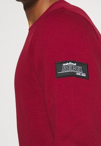 Pull rouge en maille texturée, avec un patch noir sur la manche indiquant "redefined REBEL est. 2013." Design simple et ajusté.