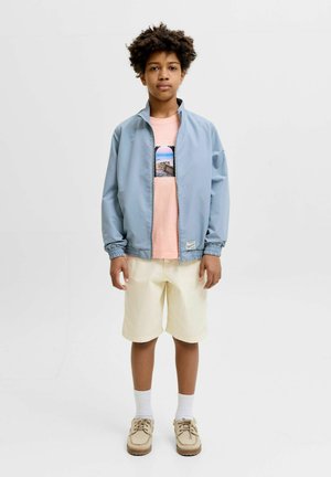 Niño con cabello rizado que lleva una chaqueta azul claro sobre una camiseta rosa con estampado, pantalones cortos crema, calcetines blancos y zapatos beige, de pie sobre un fondo blanco.