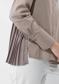 Langärmliges Hemd in hellem Taupe mit einem plisseeartigem Rückendetail, geknöpften Manschetten und glattem Stoff, kombiniert mit weißen Hosen.