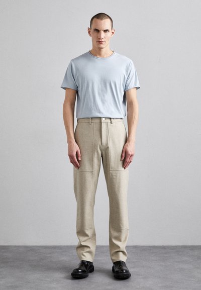 Light blue t-shirt with a round neckline paired with beige tailored pants featuring patch pockets and dark shoes, posed against a neutral background.