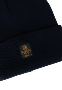 Berretto lavorato a maglia blu navy con texture a coste. Presenta una toppa quadrata nera con un'illustrazione di un cane arancione e il testo "Refugiwear".