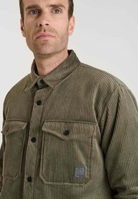 Groene corduroy overhemdjacket met twee voorzakken, zwarte knopen en een kraagachtige halslijn. Geweven stof en logo-patchdetail zichtbaar.