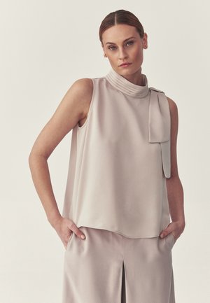 Femme portant un haut beige sans manches avec un col montant plissé et un pantalon large assorti, debout les mains dans les poches devant un fond neutre.