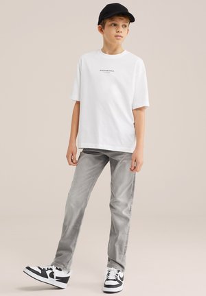 Garçon portant une casquette noire, un T-shirt blanc surdimensionné, un jean gris et des baskets noir et blanc, debout devant un fond uni.