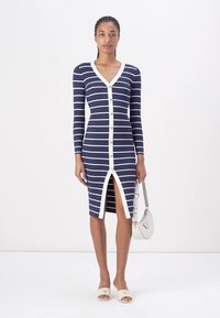 Marineblå og hvitstripet ribbet kjole med V-hals, lange ermer, frontknapper og høy splitt. Kombinert med en lys veske og sandaler.