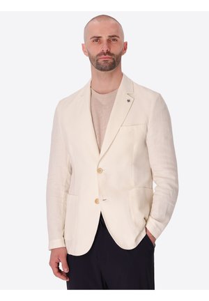 Uomo con barba corta che indossa un blazer beige su una camicia beige chiaro e pantaloni neri, in piedi con una mano in tasca su sfondo bianco.