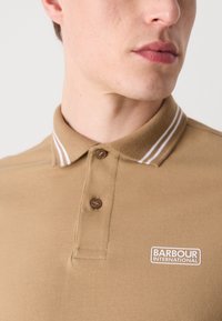 Braunes Poloshirt aus strukturiertem Baumwollstoff mit einer Knopfleiste mit zwei Knöpfen, weißen Akzentstreifen am Kragen und einem Logo-Patch auf der Brust.