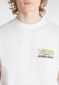 Bílá bavlněná tričko se zaobleným výstřihem. Na levém prsu se nachází žluté a černé logo "VANS DESIGN CO. custom classic".