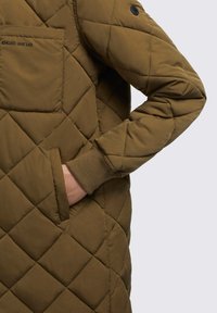 Veste matelassée vert olive avec des motifs en losanges, une poche poitrine et des poignets en tricot. Fabriquée en tissu doux et durable.