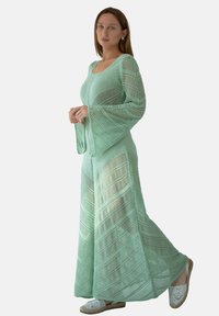 Miętowo-zielona, dzianinowa maxi sukienka z długimi, rozszerzanymi rękawami, z wzorem strukturalnym i przezroczystymi elementami. Noszona z białymi sandałami.
