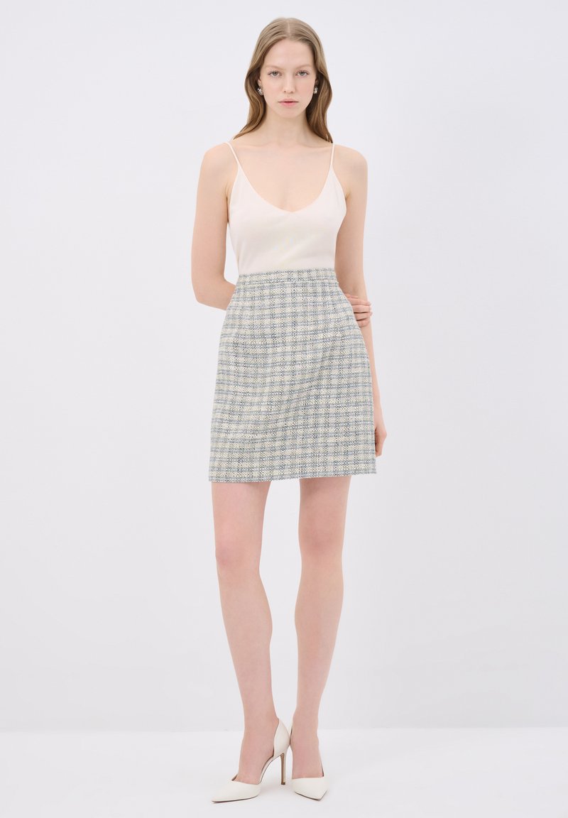 Beige tílko na tenké ramínka v kombinaci se světle modrou a bílou kostkovanou mini sukní. Modelka má na sobě bílé špičaté lodičky. Jednoduché pozadí.