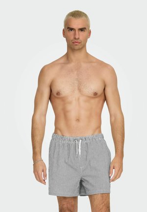 Blondyn z krótkimi włosami w szaro-białych kąpielówkach w paski, stojący na tle jasnego, jednolitego tła, zwrócony przodem.