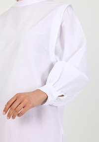 Weiße Bluse mit langen, puffartigen Ärmeln und knöpfbaren Bündchen, aus leichtem Stoff gefertigt, der ein strukturiertes, minimalistisches Design betont.