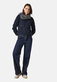 Marineblå fleecejakke med lynlås og to forlommer, parret med mørke denim bukser med vidde og et mønstret sort og hvidt tørklæde.